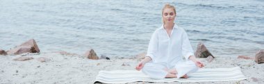 panoramic shot of peaceful blonde young woman with closed eyes practicing yoga near river  clipart