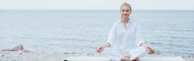 panoramic shot of peaceful blonde young woman with closed eyes practicing yoga near river  clipart