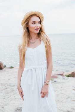 beyaz elbise ve hasır şapka ayakta iken gülümseyen güzel sarışın kız