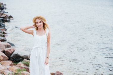 neşeli genç sarışın kadın hasır şapka dokunmadan ve deniz kenarında kamera bakarak 