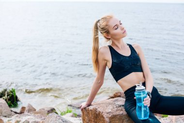 deniz kenarında taş üzerinde otururken spor şişesi tutan sarışın sporcu 