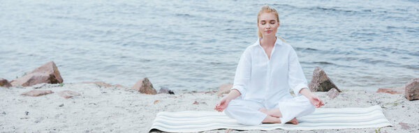 panoramic shot of peaceful blonde young woman with closed eyes practicing yoga near river 