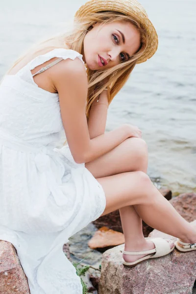 Menina Loira Atraente Tocando Chapéu Palha Enquanto Sentado Pedras — Fotografia de Stock