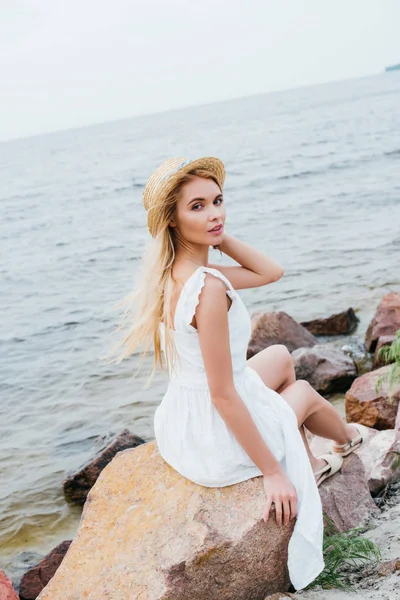 白いドレスで石の上に座っている間 美しいブロンドの女の子が麦わら帽子に触れる — ストック写真