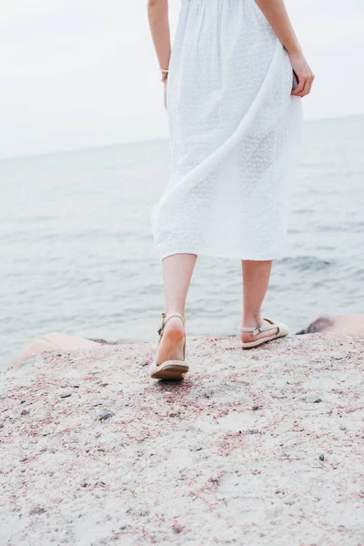 海の近くに立つ白いドレスとサンダルを着た女性のトリミングされた景色 — ストック写真