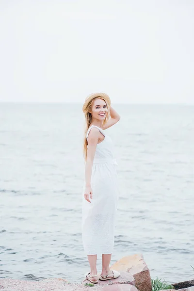 麦わら帽子に触れ 海の近くに白いドレスを着て立っている間 カメラを見ている笑顔のブロンドの若い女性 — ストック写真