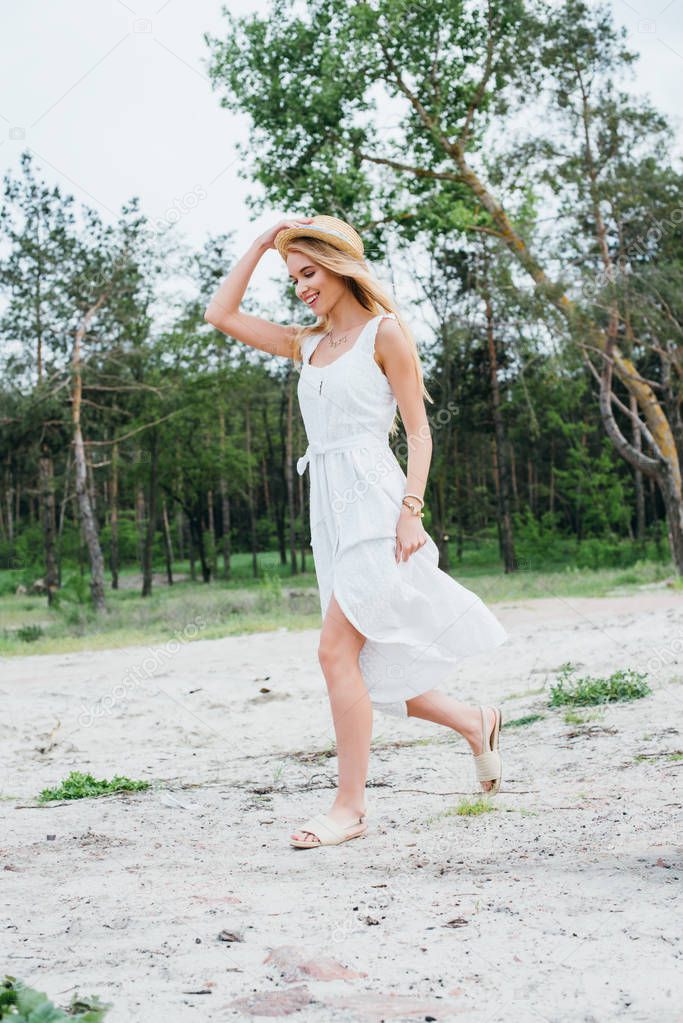 happy blonde girl smiling while touching straw hat and walking near green trees 