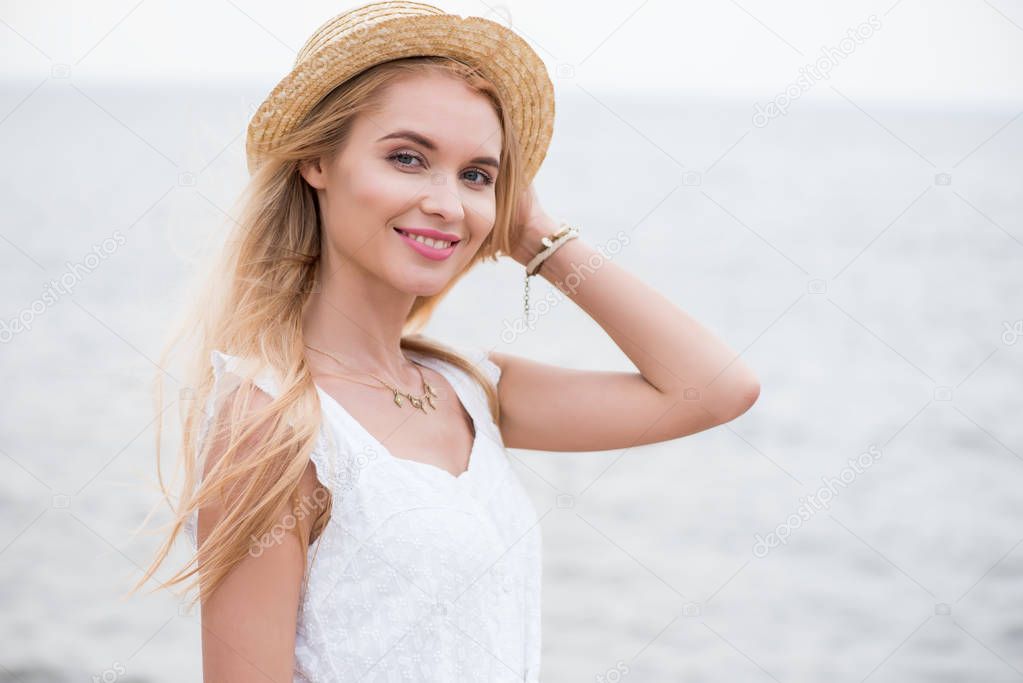 beautiful blonde young woman looking at camera and touching straw hat near sea 