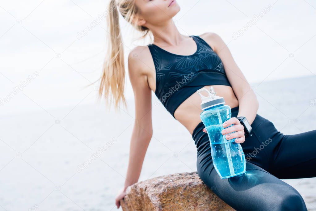 cropped view of sportswoman holding sport bottle while sitting on stone near sea 