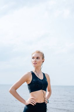 atletik genç kadın kalça elleri ile ayakta ve deniz kenarında kulaklıkmüzik dinleme