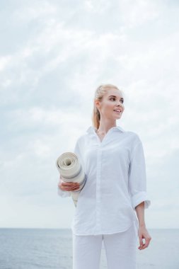 low angle view view of cheerful young blonde woman standing near sea and holding yoga mat  clipart