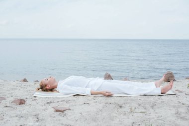 attractive blonde woman with closed eyes meditating while lying on yoga mat near sea  clipart