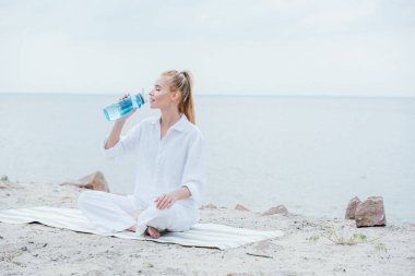 çekici kadın yoga mat üzerinde oturan ve su içerken spor şişesi tutarak 