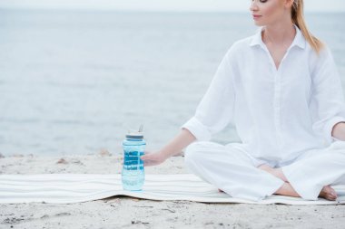 spor şişesi yakınında yoga mat üzerinde oturan neşeli kadın kırpılmış görünümü 