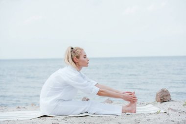 deniz kenarında yoga minderi üzerinde otururken esneme çekici kadın yan görünümü 