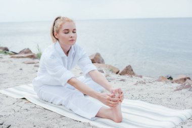 deniz kenarında yoga minderinde otururken uzanan kapalı gözlü çekici kadın 