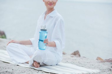 cropped view of cheerful woman sitting on yoga mat and holding sport bottle near sea  clipart