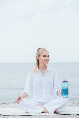 neşeli sarışın kadın spor şişesi tutan ve deniz kenarında yoga mat oturuyor 