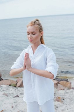 attractive young woman with closed eyes and praying hands near sea  clipart