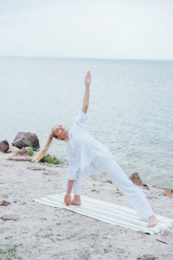 Gözleri kapalı genç bir kadın deniz kenarındaki yoga minderinde yoga yapıyor. 