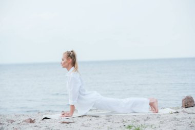 yoga mat üzerinde nehir yakınında yoga yapan sarışın genç kadının yan görünümü