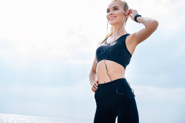 low angle view of cheerful athletic girl listening music while standing with hand on hip 