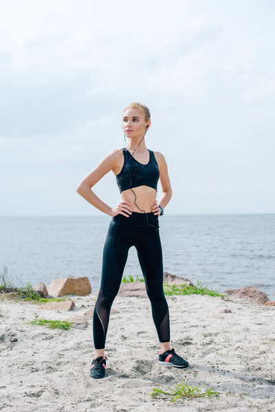 athletic girl standing with hands on hips and listening music in earphones near sea