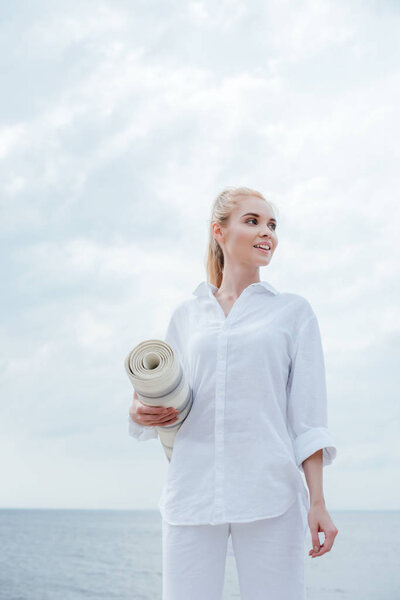 low angle view view of cheerful young blonde woman standing near sea and holding yoga mat 