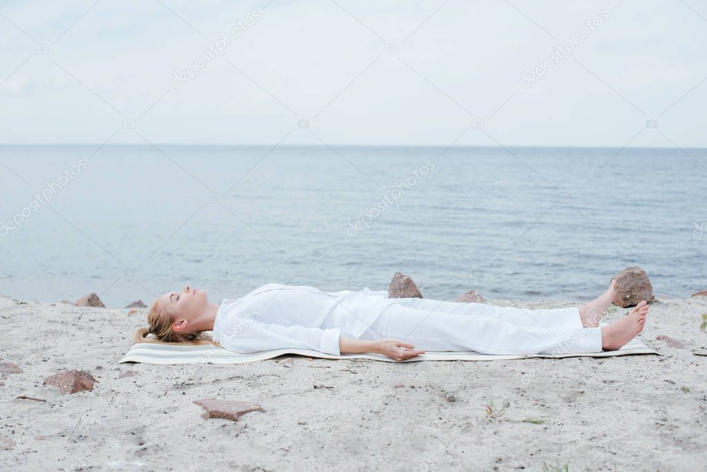 attractive blonde woman with closed eyes meditating while lying on yoga mat near sea 