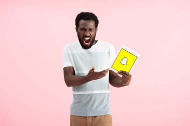 KYIV, UKRAINE - MAY 17, 2019: emotional african american man shouting and showing digital tablet with Snapchat app, isolated on pink clipart