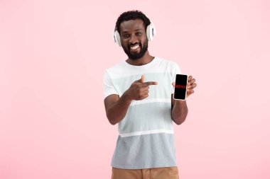 Kiev, Ukrayna - 17 Mayıs 2019: Afro-Amerikan adam kulaklıkla müzik dinliyor ve netflix uygulamasıyla akıllı telefona işaret ediyor, pembeye izole 