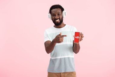 Kiev, Ukrayna - 17 Mayıs 2019: Afro-Amerikan adam kulaklıkla müzik dinliyor ve youtube uygulamasıyla akıllı telefona işaret ediyor, pembeye izole 