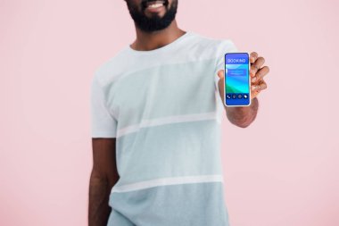 rezervasyon uygulaması ile akıllı telefon gösteren Afrikalı-Amerikalı adam kırpılmış görünümü, pembe izole 