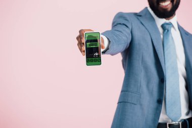 rezervasyon uygulaması ile akıllı telefon gösteren takım elbiseli Afrikalı Amerikalı işadamının kırpılmış görünümü, pembe izole 