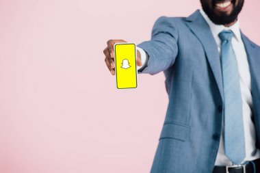 Kiev, Ukrayna - 17 Mayıs 2019: Snapchat uygulaması ile akıllı telefon gösteren takım elbiseli Afrikalı Amerikalı işadamının kırpılmış görünümü, pembe izole 