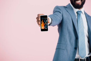 infografik uygulaması ile akıllı telefon gösteren takım elbiseli Afrikalı Amerikalı işadamının kırpılmış görünümü, pembe izole 