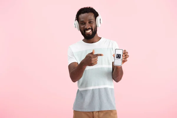乌克兰 2019年5月17日 非洲裔美国男子听音乐与耳机 并指向智能手机与优步应用程序 隔离在粉红色 — 图库照片