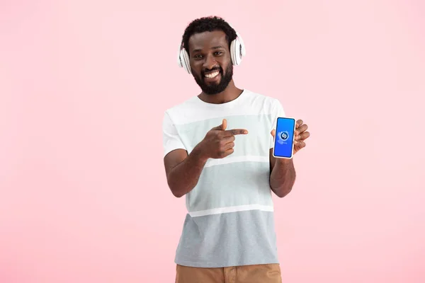 乌克兰 2019年5月17日 非洲裔美国男子听音乐与耳机 并指向智能手机与Shazam应用程序 隔离在粉红色 — 图库照片