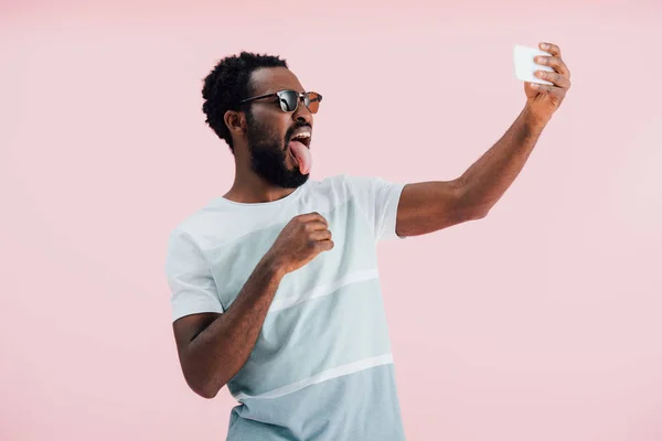 サングラスをかけたアフリカ系アメリカ人男性が舌を出し スマートフォンで自分撮りを取り ピンクで孤立 — ストック写真