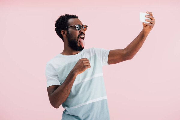 african american man in sunglasses sticking tongue out and taking selfie on smartphone, isolated on pink 