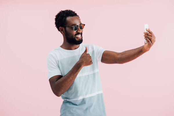 african american man in sunglasses with thumb up taking selfie on smartphone, isolated on pink 