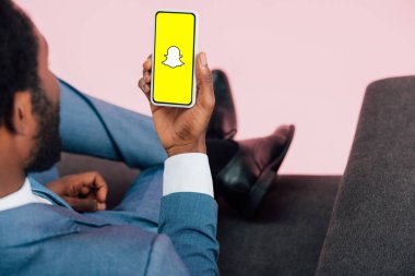 KYIV, UKRAINE - MAY 17, 2019: cropped view of african american businessman sitting on armchair and showing smartphone with Snapchat app, isolated on pink clipart