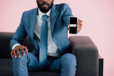 Kiev, Ukrayna - 17 Mayıs 2019: Afro-Amerikan işadamının koltukta oturan ve hbo uygulaması ile akıllı telefon gösteren, pembe izole görünümü kırpılmış