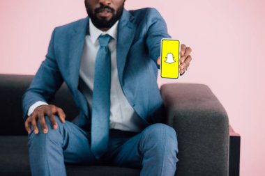 KYIV, UKRAINE - MAY 17, 2019: cropped view of african american businessman sitting on armchair and showing smartphone with Snapchat app, isolated on pink clipart