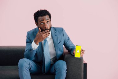 KYIV, UKRAINE - MAY 17, 2019: shocked african american businessman in suit sitting on armchair and showing smartphone with Snapchat app, isolated on pink clipart
