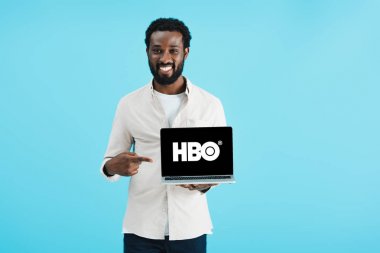 KYIV, UKRAINE - MAY 17, 2019: smiling african american man pointing at laptop with HBO website, isolated on blue clipart