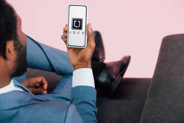 キエフ ウクライナ 2019年5月17日 アームチェアに座って ピンクに隔離されたUberアプリでスマートフォンを示すアフリカ系アメリカ人ビジネスマンのトリミングビュー — ストック写真