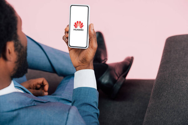 KYIV, UKRAINE - MAY 17, 2019: cropped view of african american businessman sitting on armchair and showing smartphone with huawei app, isolated on pink
