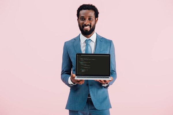 КИЕВ, УКРАИНА - 17 мая 2019 года: улыбающийся африканский бизнесмен, показывающий ноутбук с HTML-кодом, изолированный на розовом
