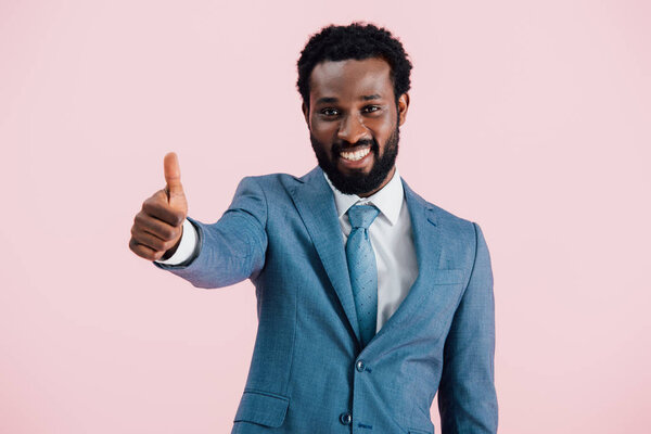 smiling african american businessman in suit showing thumb up, isolated on pink